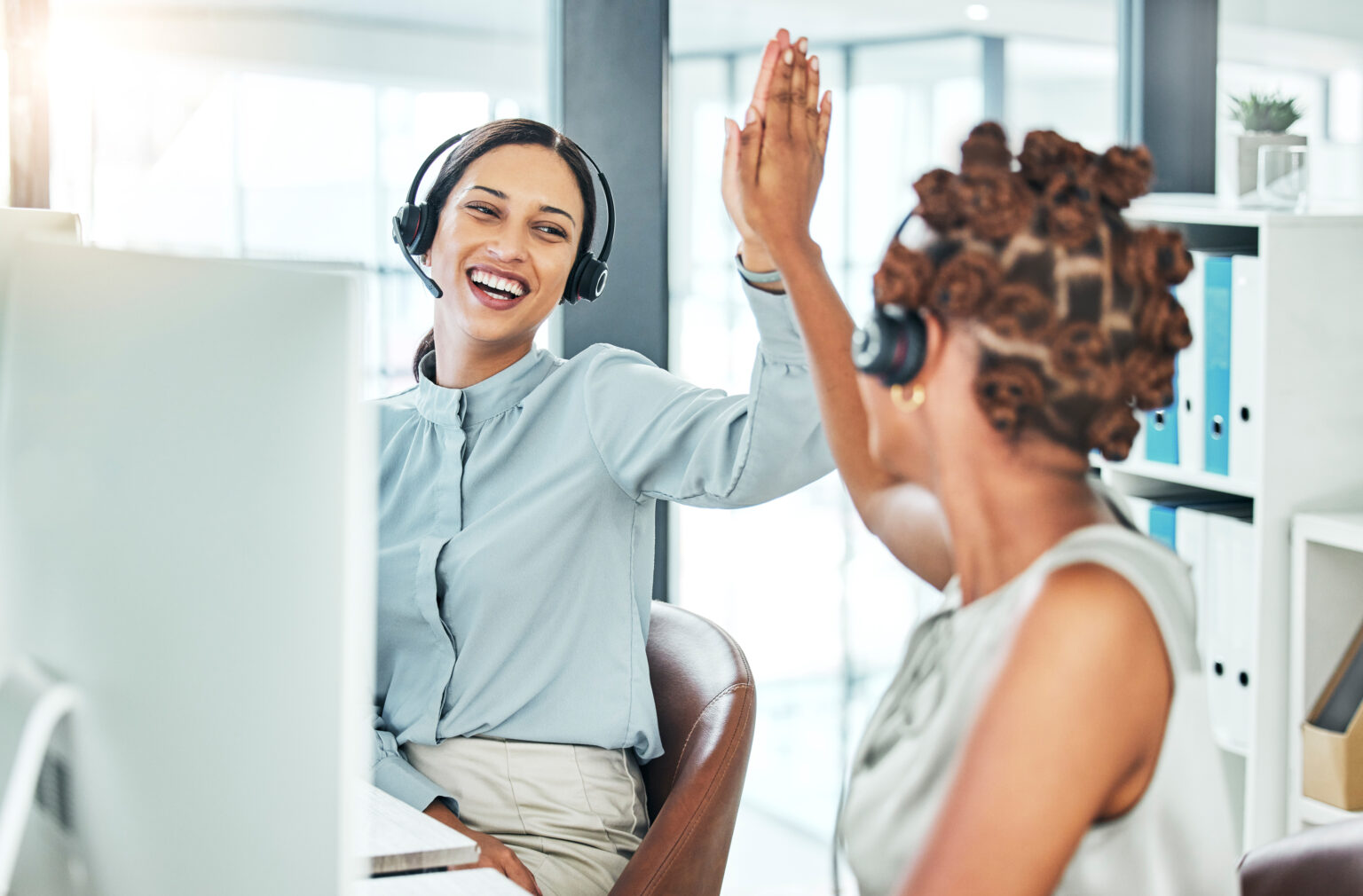 High five, excited and celebrating call center agents with computers happy with a successful deal, promotion and sale. Smiling, cheering and motivated contact us customer service operators in office.